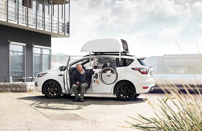 Mann, der in einem Turny Evo-Sessellift sitzt, schwenkte aus dem Auto mit einer Rollstuhl-Dachbox des Stuhls Topper