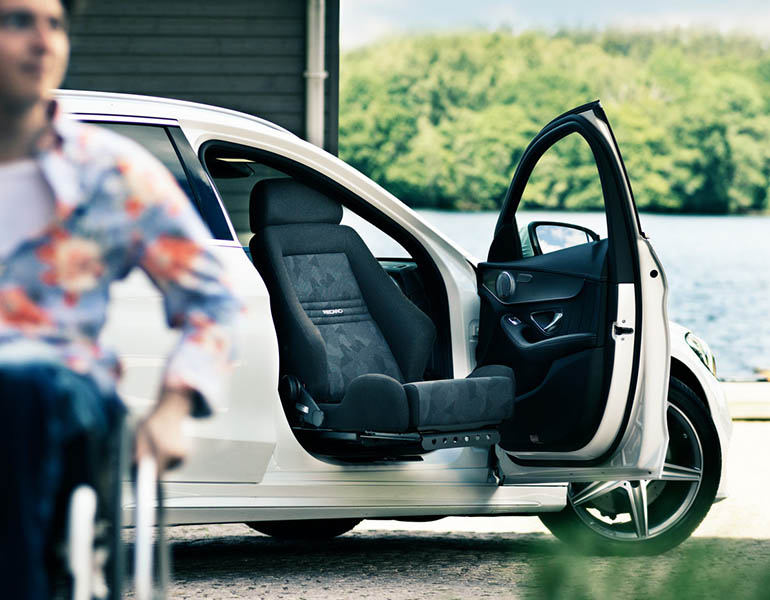 Une voiture blanche avec un siège Recaro pivotant sur le siège du passager avant. Un homme flou se dirige vers le spectateur en fauteuil roulant.