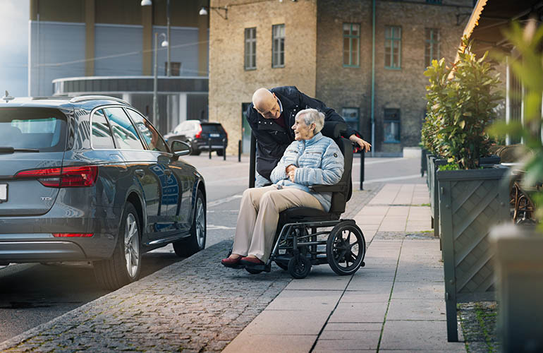 An elderly couple talking to each other. The woman is seated in a wheelchair and the man is standing behind her, leaning in to listen. 