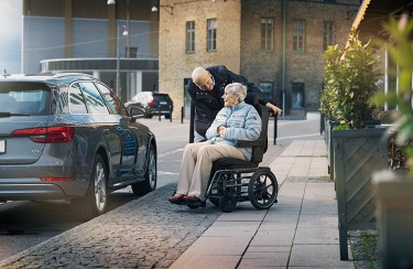 Ein älteres Ehepaar spricht miteinander. Die Frau sitzt im Rollstuhl, der Mann steht hinter ihr und beugt sich vor, um zuzuhören.