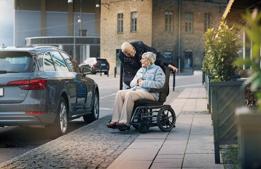 An elderly couple talking to each other. The woman is seated in a wheelchair and the man is standing behind her, leaning in to listen. 