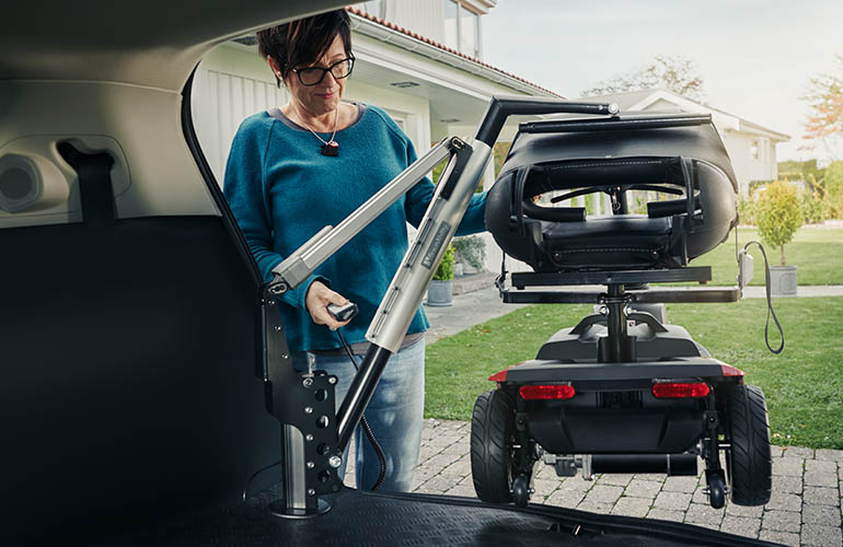 The Carolift 100 and a mobility scooter, shown from the inside of the car boot, and a woman standing behind it with one hand on the scooter and the other holding a remote control.
