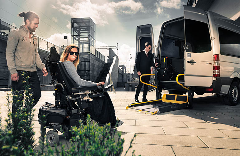 Frau im Elektrorollstuhl steht kurz vor dem Einstieg in ein Transportfahrzeug mit Rollstuhllift