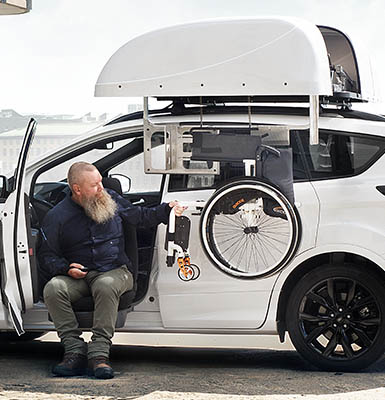 A man in the front seat of a car using a roof top box wheelchair hoist. 