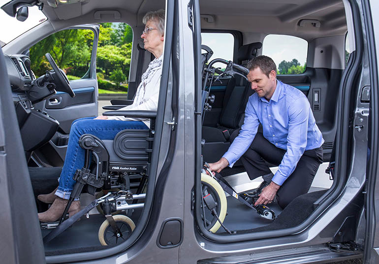 Seitenansicht eines Mannes, der eine ältere Frau in einem Rollstuhl für den Transport in einem Fahrzeug sichert.