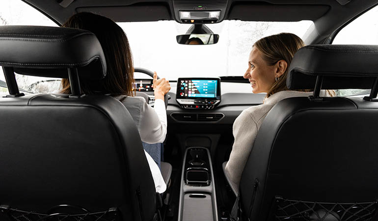 Two young women in the front seat of a car. The passenger is looking at the driver. 