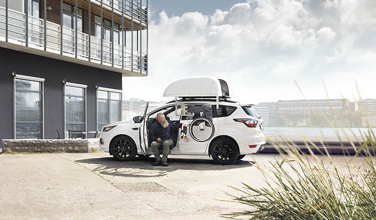 A man sitting in the front seat of a parked car facing out of the car retreiving his wheelchair from a rooftop box hoist