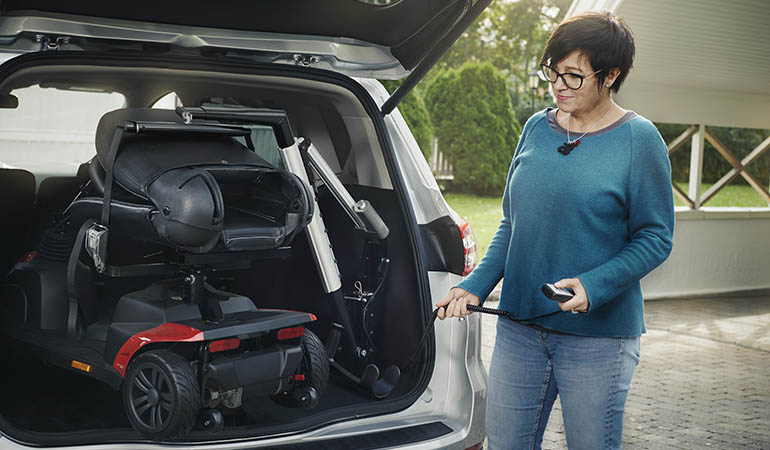 A woman standing next to the boot of a car using a remote control to load her scooter into the car using a boot hoist.