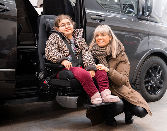 Girl sitting in a specially adapted seat attached to the Turny Evo seat lift outside of a car and her mother crouching next to her