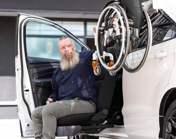 Man sitting in a car seat attached to the Turny Evo seat lift outside of a car retrieving his wheelchair from the Chair Topper roof top box.