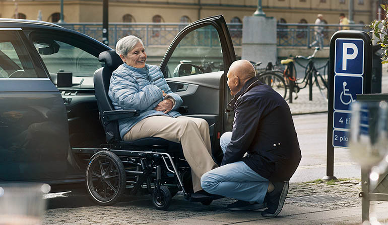 Une femme assise dans un Carony est assistée dans la voiture par un homme