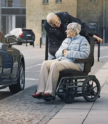 Mann, der leicht hinter Frau auf Rollstuhl steht und sie anlächelt