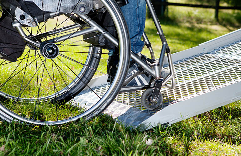 The bottom part of a wheelchair rolling onto a ramp deployed on grass. 