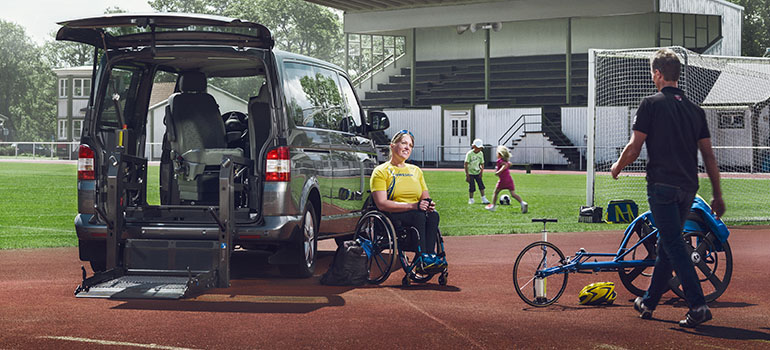 In einem Outdoor-Sportplatz sitzt Frau auf einem Rollstuhl neben einem geparkten Auto