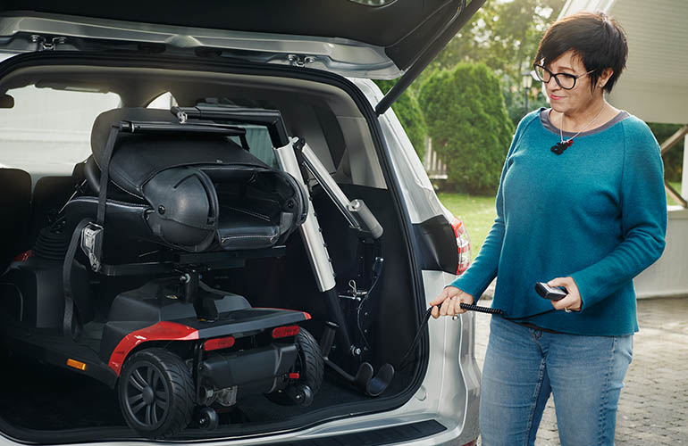 Le coffre d'une voiture avec un scooter de mobilité attaché à un Carolift 100 à l'intérieur, et une femme debout à côté de la voiture tenant une télécommande.