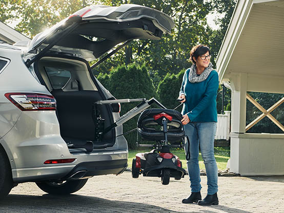 A woman standing on the driveway by the boot of a parked car, using the Carolift 100 with a remote control to fetch her mobility scooter. 