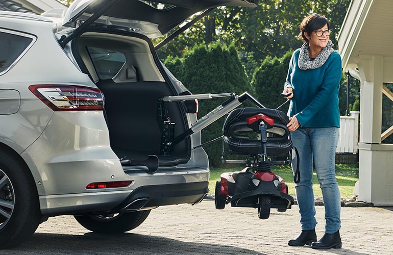 A woman standing on the driveway by the boot of a parked car, using the Carolift 100 with a remote control to fetch her mobility scooter. 