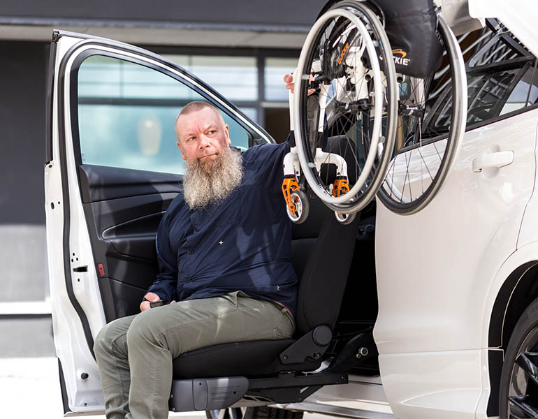 A man seated in a car seat lift, reaching for a wheelchair hanging down from the car's rooftop box.