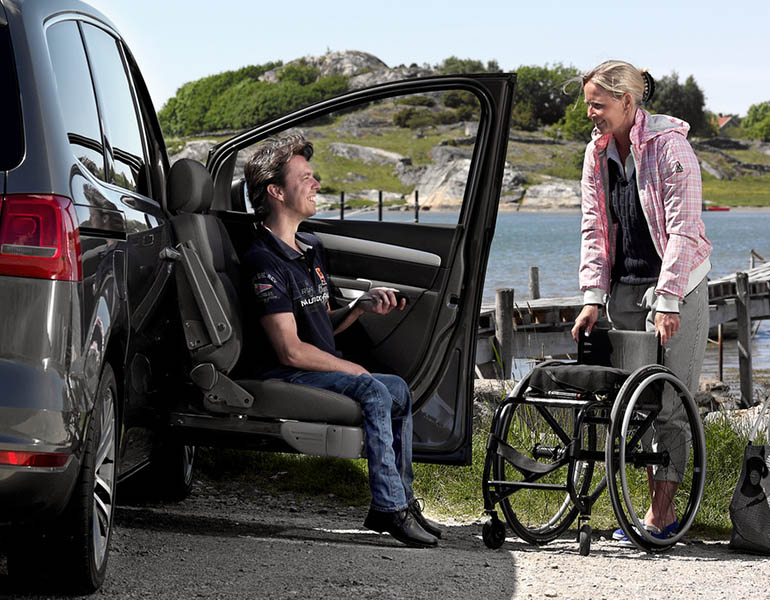 A woman brings a wheelchair to a man seated in a seat lift on the outside of a car.