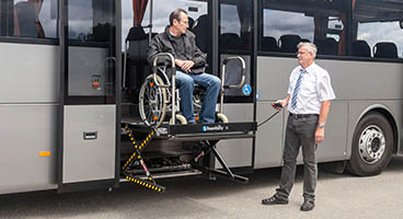 Man assisting a man on a wheelchair off of a bus with a lift
