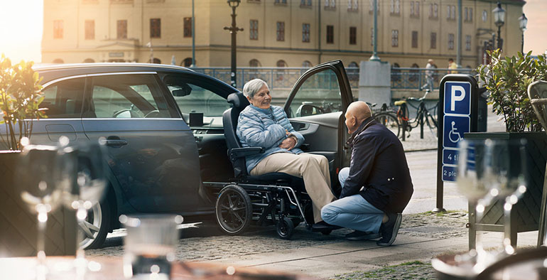 Homme âgé aidant une femme âgée à sortir d'une voiture à l'aide d'un fauteuil roulant de transfert.
