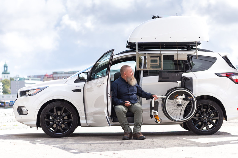 Man seated in the seat lift of a car and handling a roof rack wheelchair hoist. 