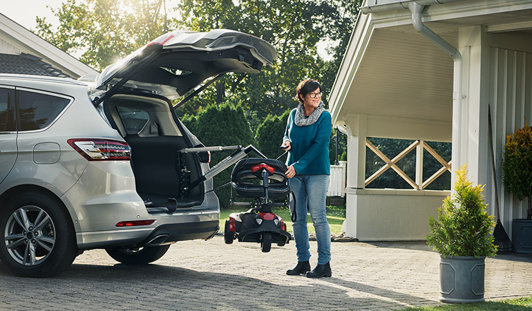 Woman standing at the boot of a car using a mobility hoist to retrieve her scooter
