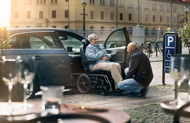 Frau in einem Carony-Rollstuhl neben einem Auto mit offener Beifahrertür und kniendem Mann