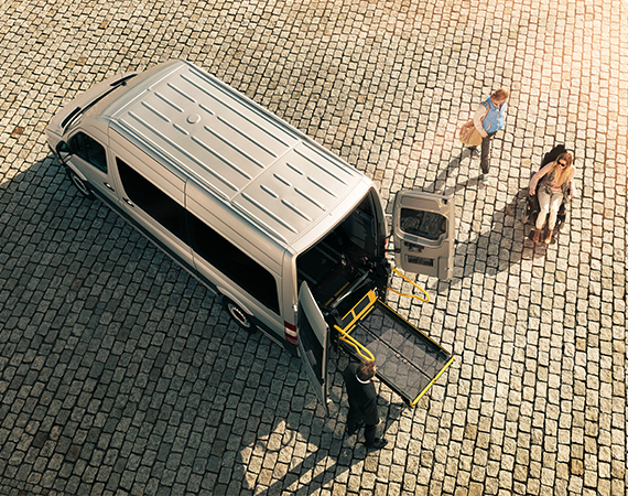 A person in a wheelchair and their carer approaching a commercial vehicle with a ramp folded out of the back and a driver standing next to it.