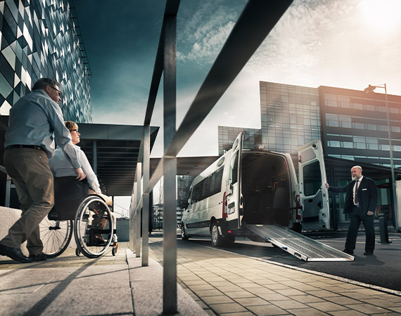 A person in a wheelchair and their carer approaching a commercial vehicle with a ramp folded out of the back and a driver standing next to it. 