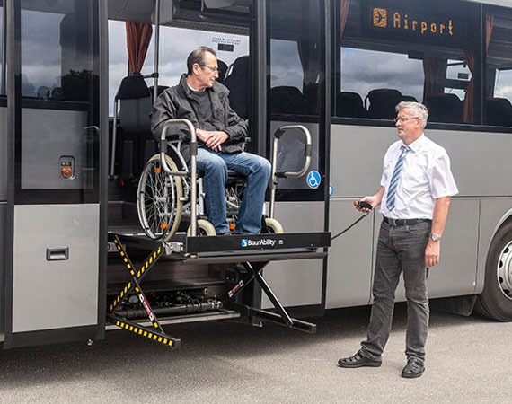 A person in a wheelchair and their carer approaching a commercial vehicle with a ramp folded out of the back and a driver standing next to it. 