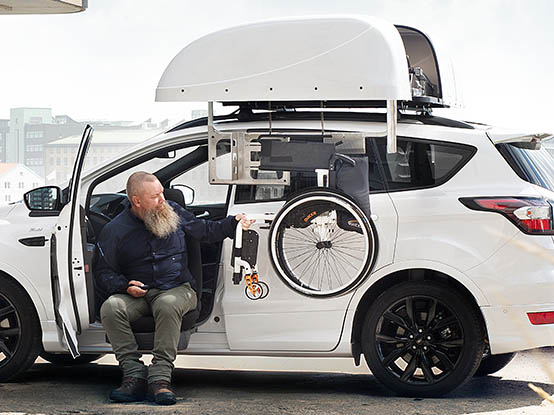 Man sitting in a white car retrieving a wheelchair from the top of the car with a chair topper