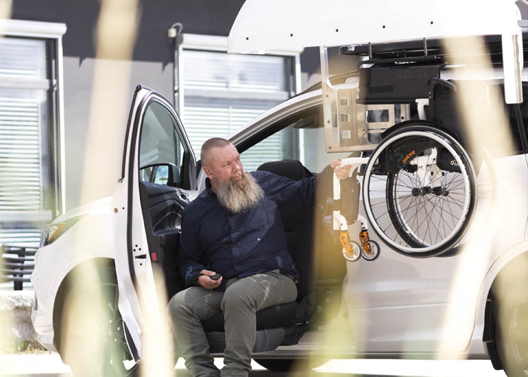 Man sitting in a car retrieving a wheelchair from the top of the car with a chair topper