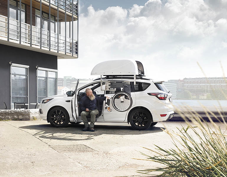 Man sitting in a white car retrieving a wheelchair from the top of the car with a chair topper