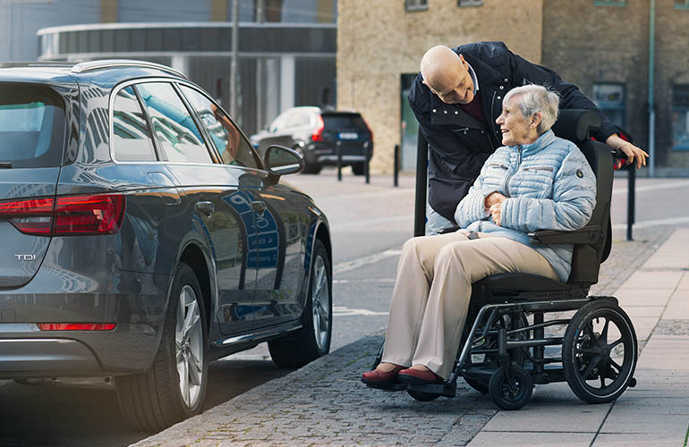 Woman in a Carony wheelchair with a man holding it next to a car