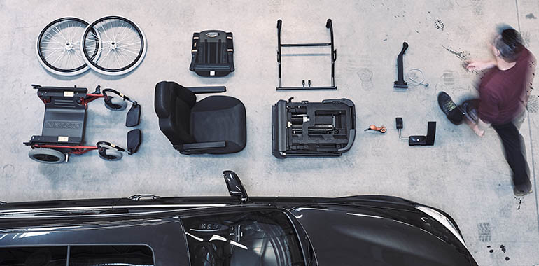 A man walking by parts of a transfer wheelchair laid out next to a car. 