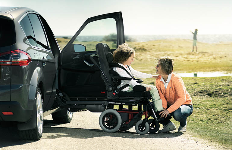 Child in a Carony wheelchair next to a car looking at her mother